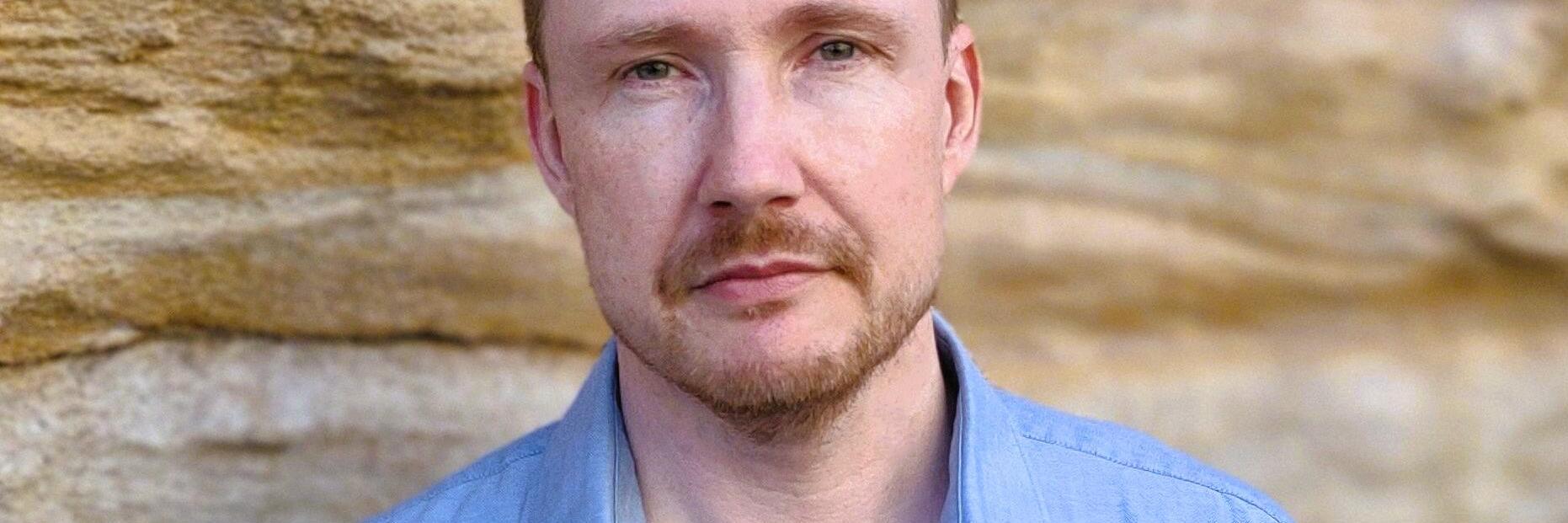 Photograph of a man with light skin, short hair and a blue shirt standing in front of a sandstone wall. 