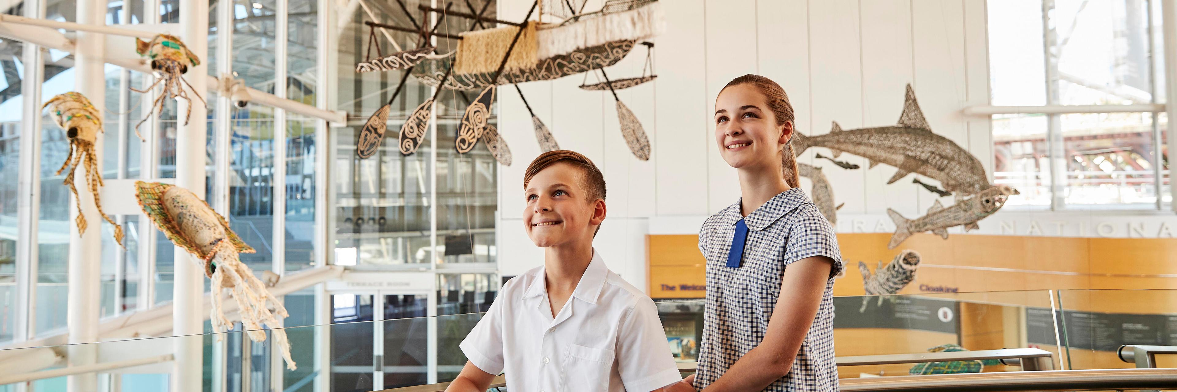 2 School kids exploring the museum, stop to look at the ghost net sculptures suspended in the foyer.