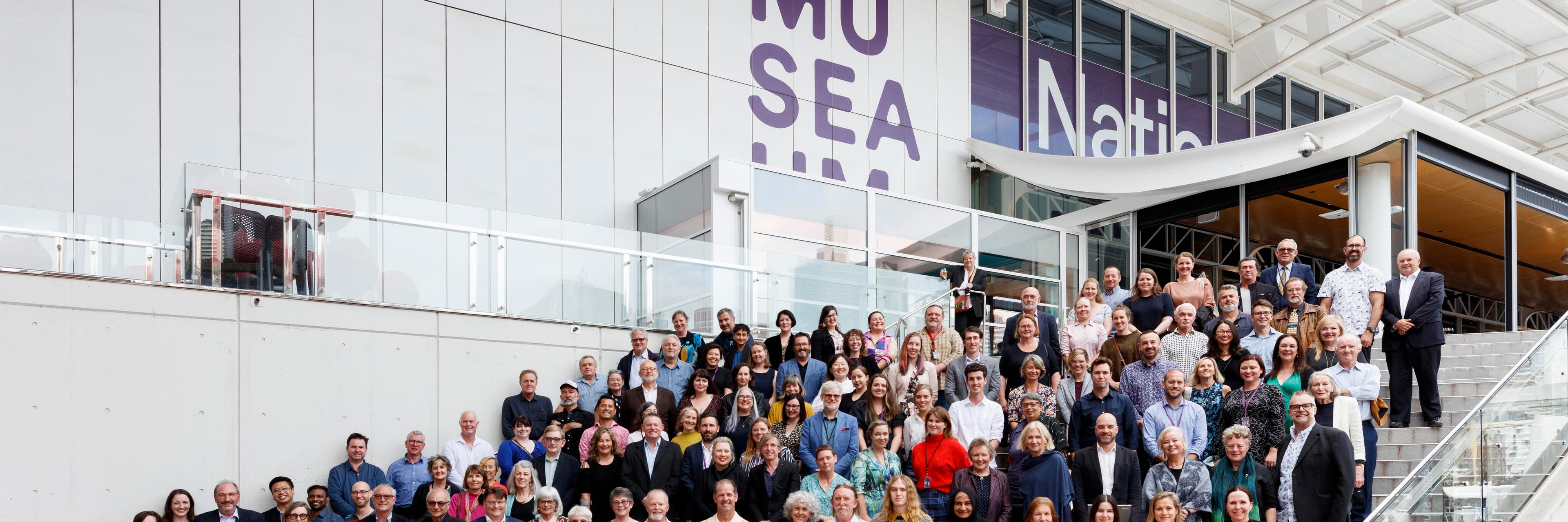 large group of people standing on wide steps in front of a white building with the museum logo on it. 