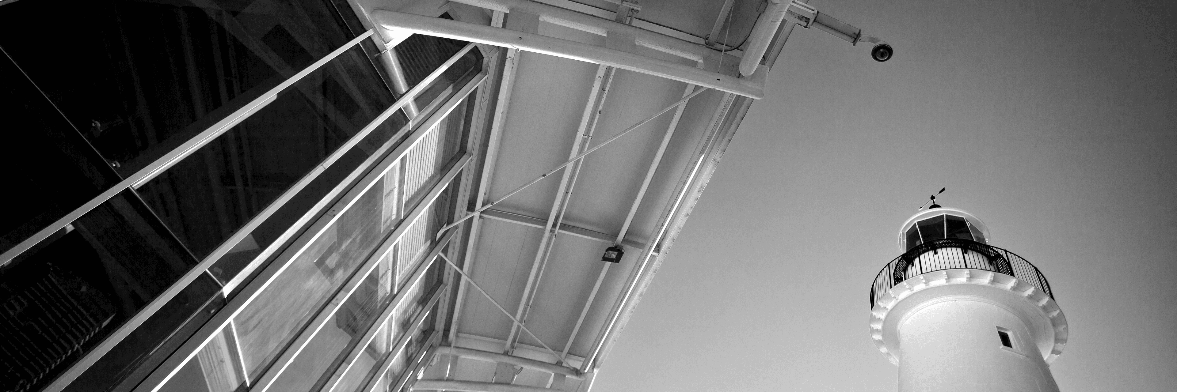 Black and white drematic photo looking up at the edge of a modern building and a lighthouse. 