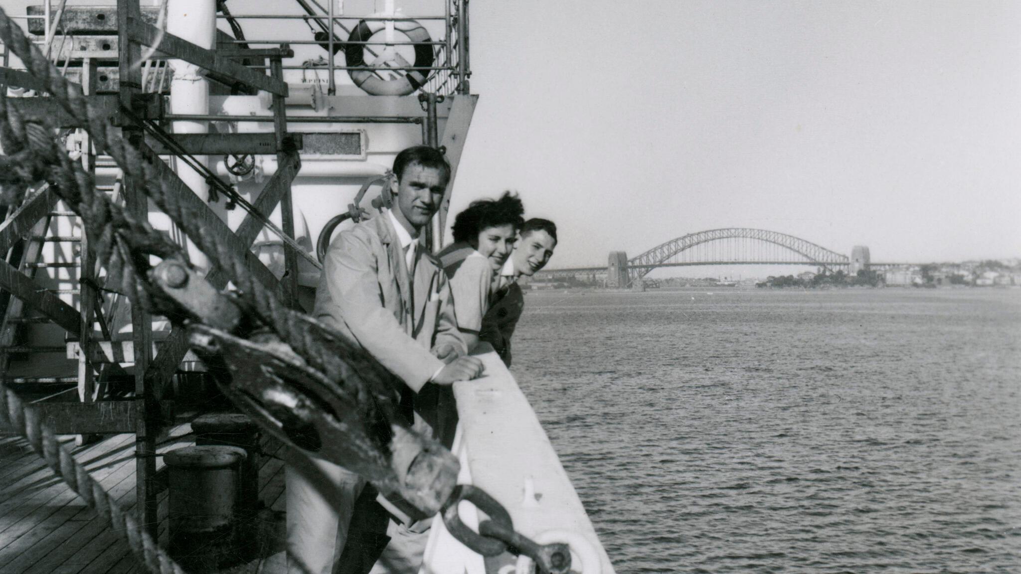 Black and White photograph, three passengers on the deck of migrant ship MV NAPOLI as it moves up Sydney Harbour. The Harbour Bridge can be seen in the background.