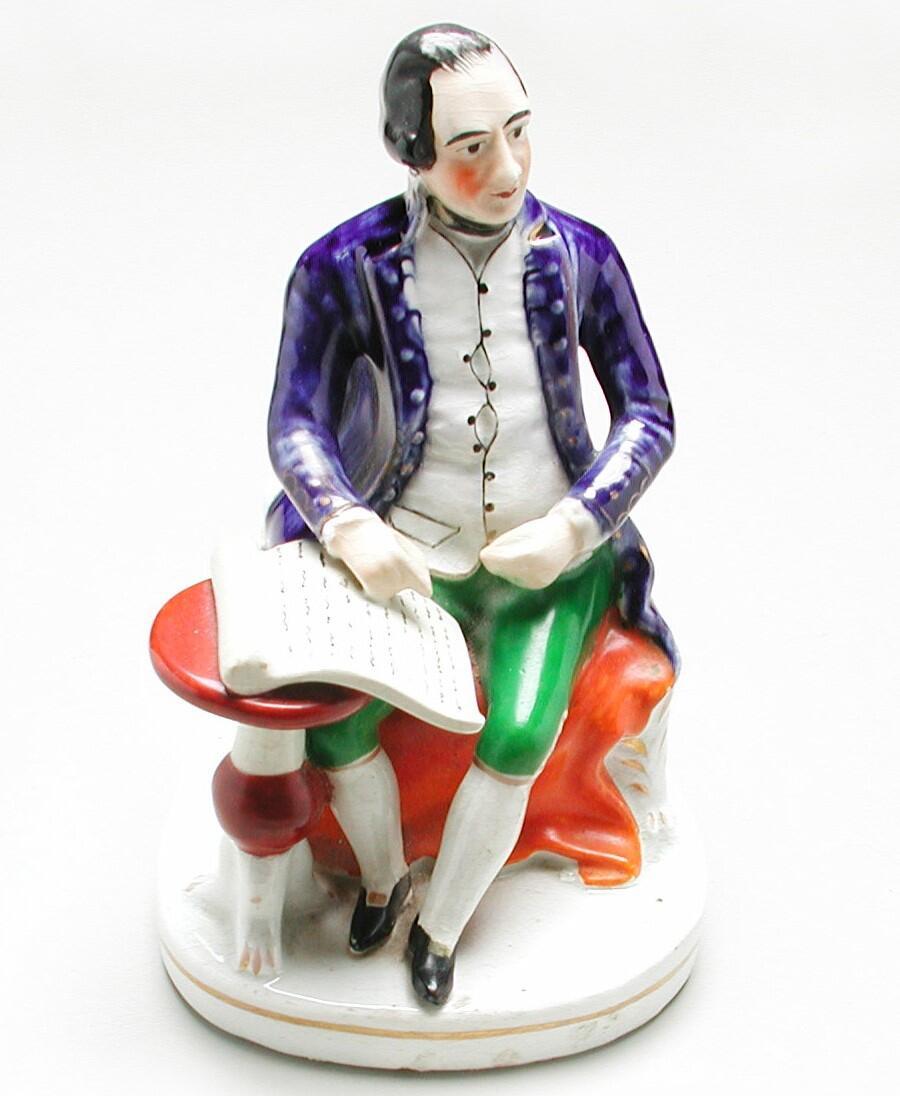 Figurine shows Cook seated, bare-headed, wearing captain's full dress uniform:  blue coat and green pantaloons. To his right is a round table upon which is a manuscript.