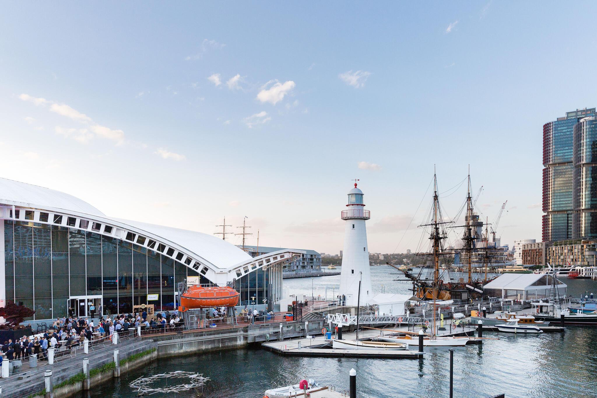 The museum precinct filled with crowds during a corporate event. museum building on the left, with lighthouse and tall ship on the right.