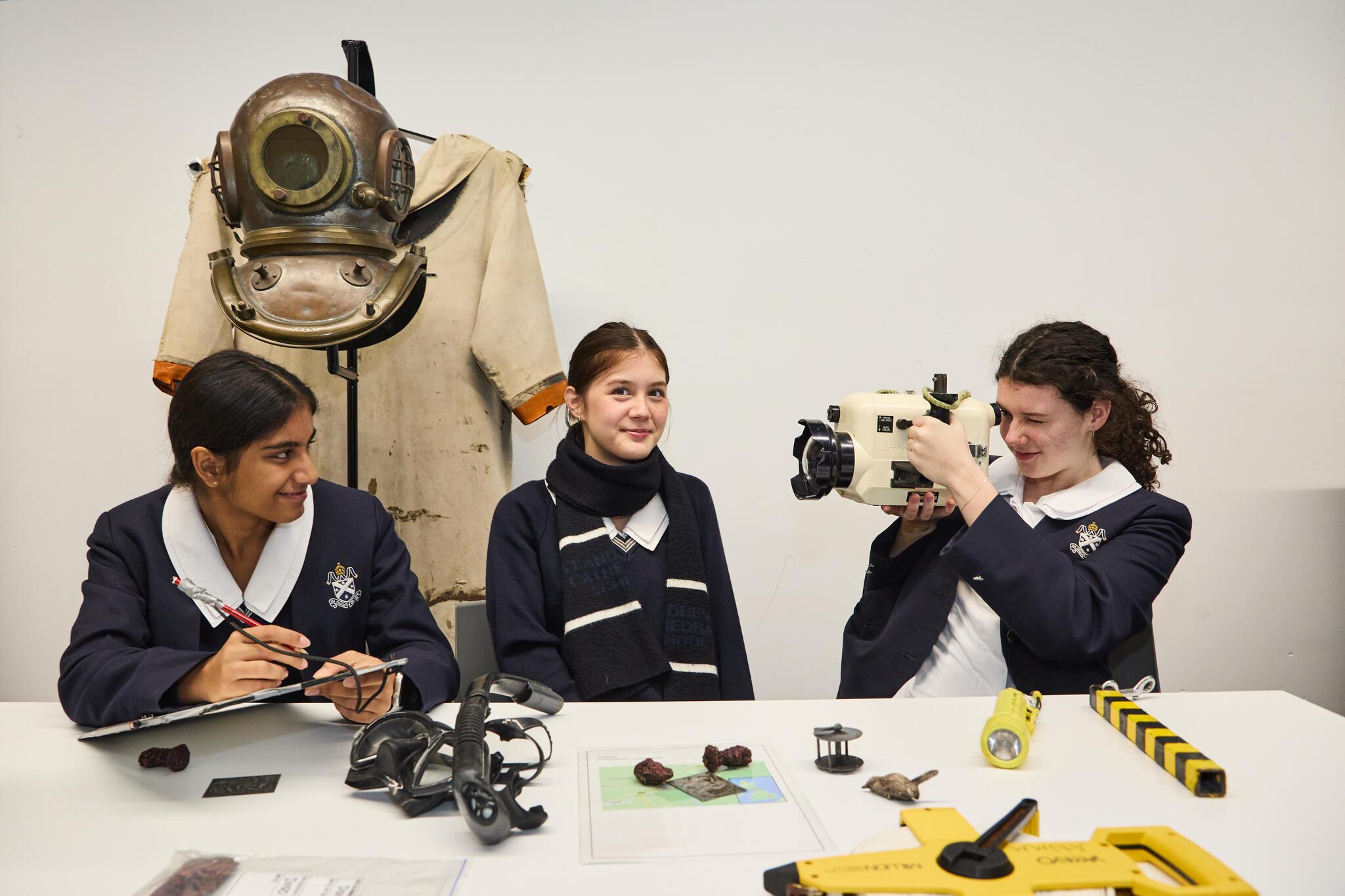 3 students in a white classroom looking at various peoices of maritime archaeology equiptment. 