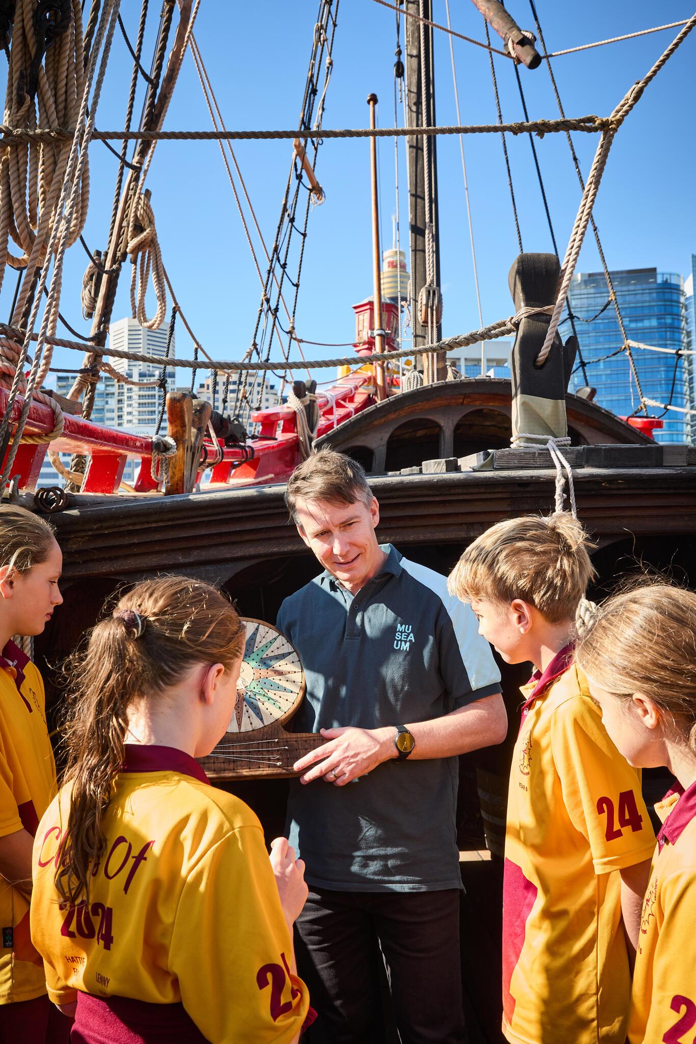 A male museum educator stands with 4 students around him onboard the Duyfken