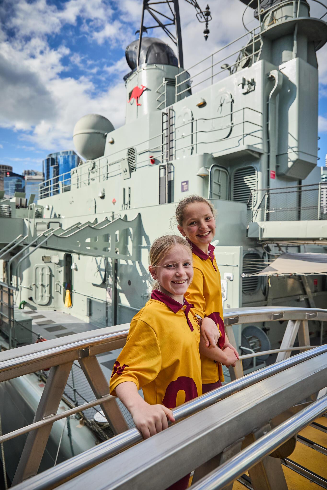 Two firls in a yellow and red school uniform on a gangway with the navy vessel vampire behind them.