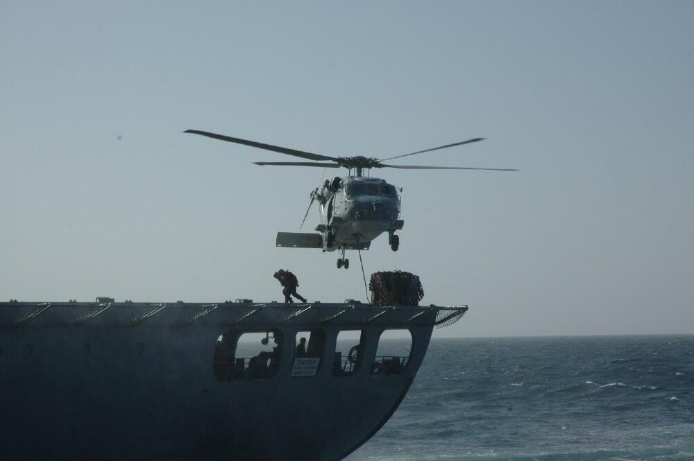 A helicopter taking off from the back of a ship, men are moving a pile of cargo beneath it. 