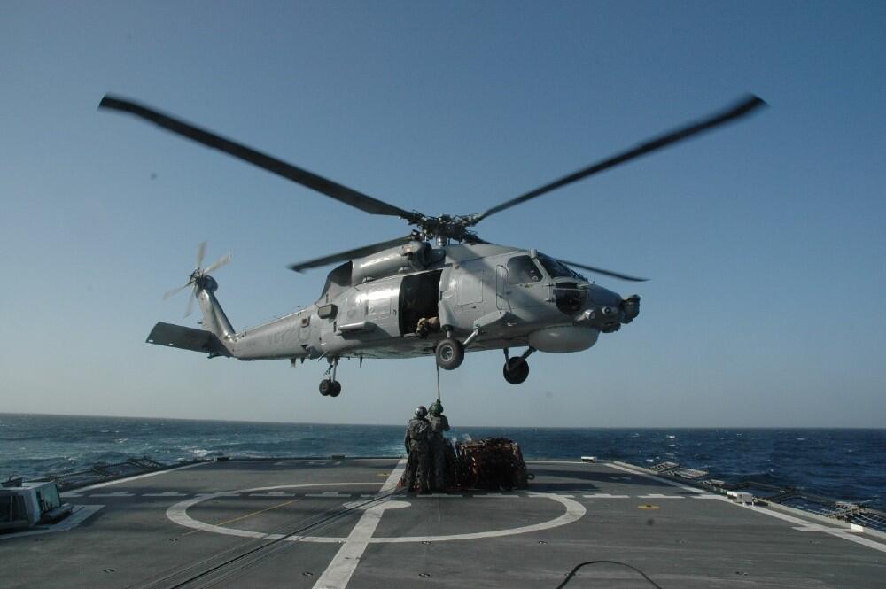 A grey navy helicopter hovering over the deck of a ship, a pile of cargo on the deck beneath it.