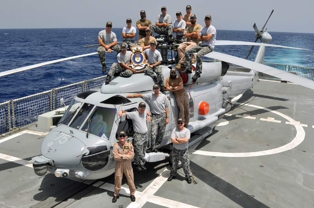 Group of military personal posing on and around a grey helicopter on the back of a ship with the ocean in the background.