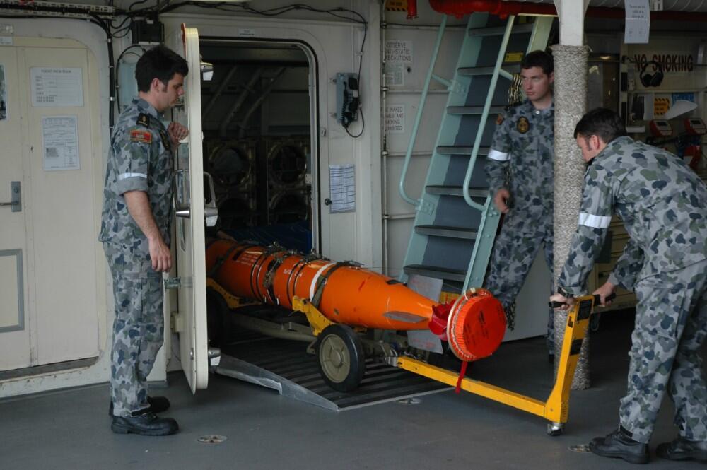 Three men in milirary uniform moving a large orange torpedo