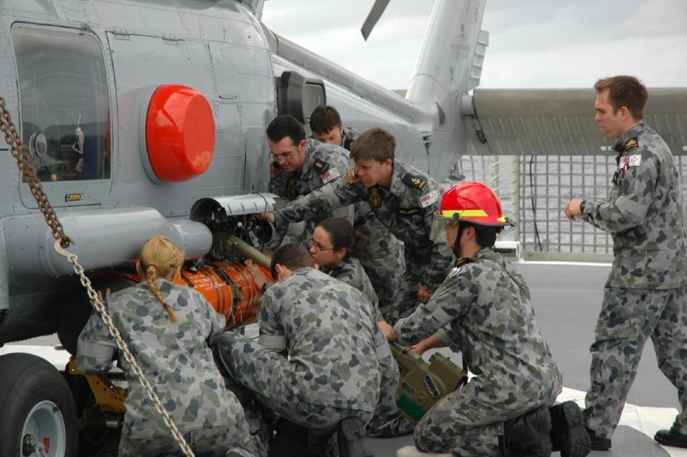 A group of military personal crowding around the side of a grey helicopter, working together to attatch a bright orange torpedo. 