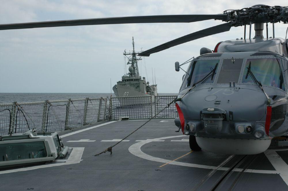 A grey military helicopter on a ship's deck, with another navy ship in the background.