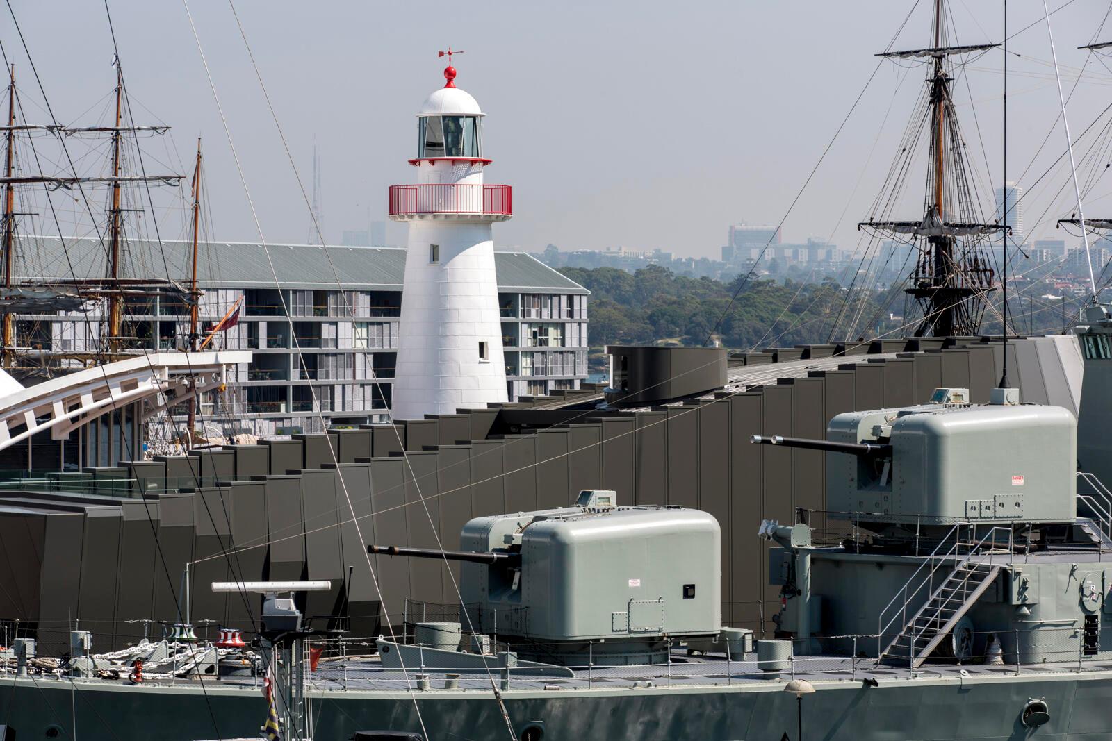 The guns of the VAMPIRE destroyer moored alongside dark grey modern building. The Cape Bowling Green Lighthouse punctuates the space behind.