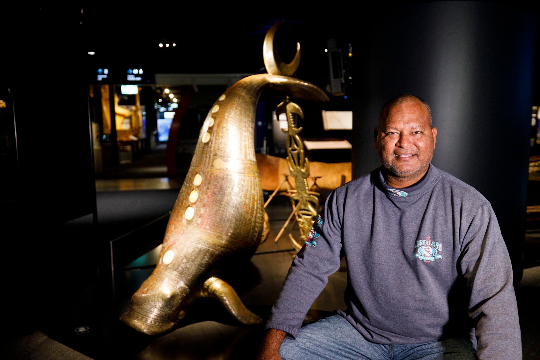 Photo of Alick Tipoti a Torres Strait Islander artist seated in front of a large bronze sculpture of a Dugong. 