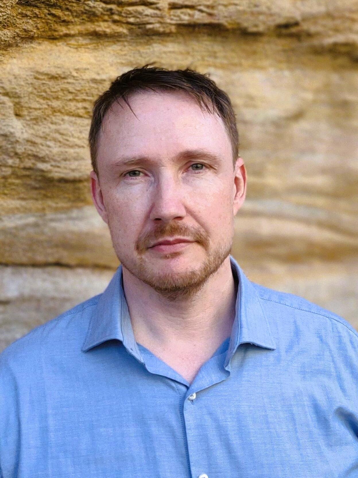 Photograph of a man with light skin, short hair and a blue shirt standing in front of a sandstone wall. 