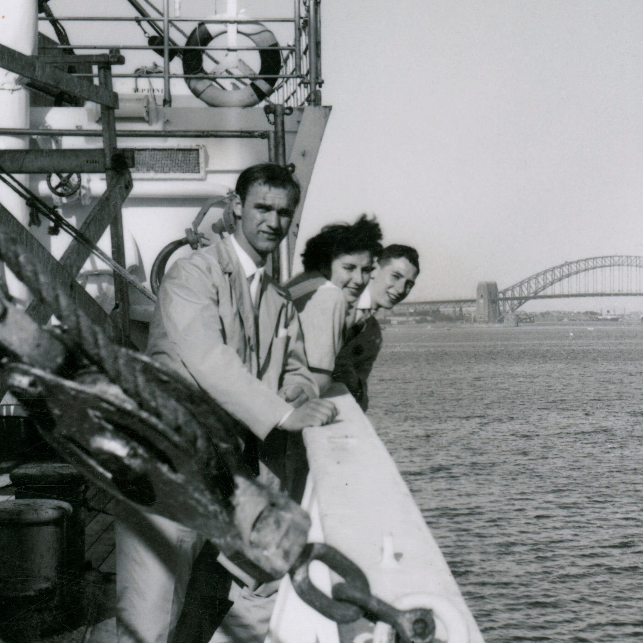 Black and White photograph, three passengers on the deck of migrant ship MV NAPOLI as it moves up Sydney Harbour. The Harbour Bridge can be seen in the background.