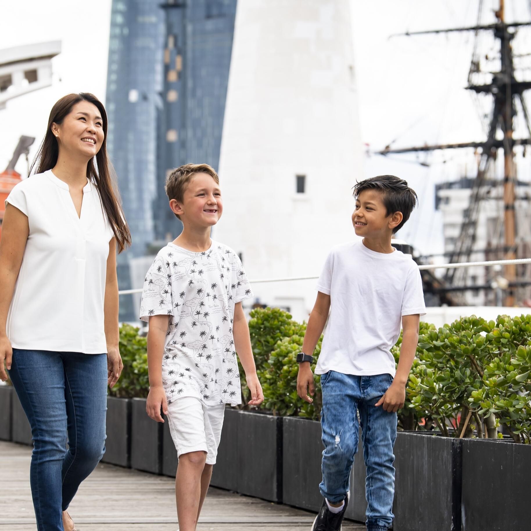 Photo of a women and two boys walking down a boardwalk, with shrubs, a lighthouse, tall ship masts and orange lifeboat in the background. 