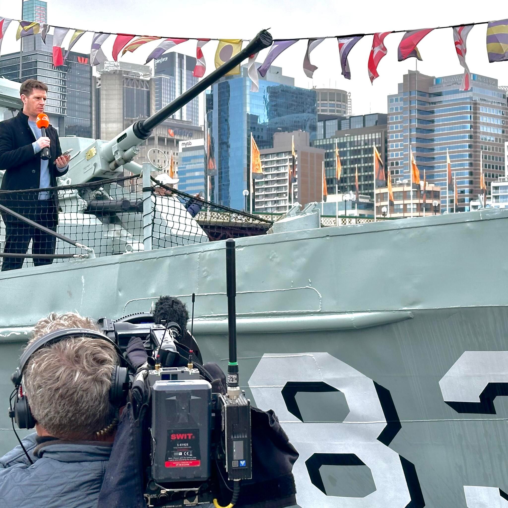 Photo showing behind the scenes off a TV shoot on a navy vessel, with a camera man in the foreground, and a male presenter standing on the boat behind.