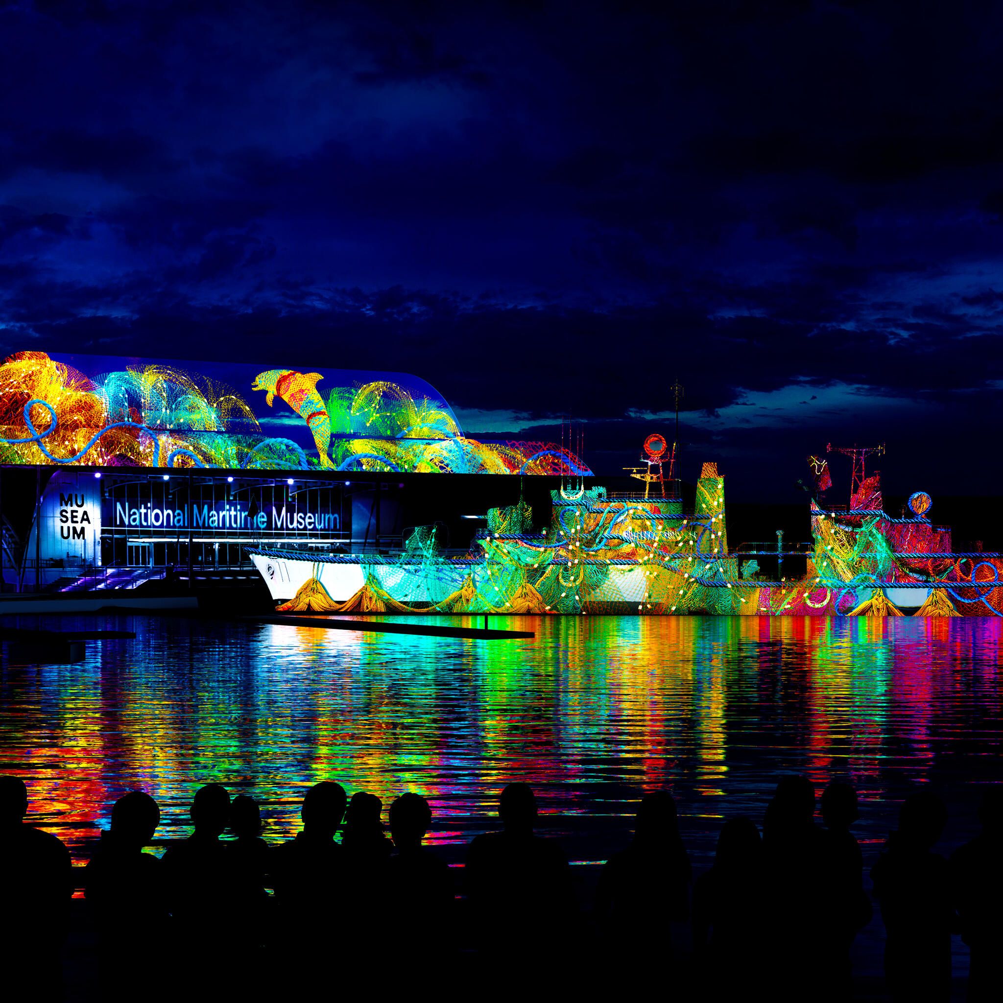 photo of colourful projection on the side of the museum and navy destroyer at night, with multicoloured reflections on the water. 