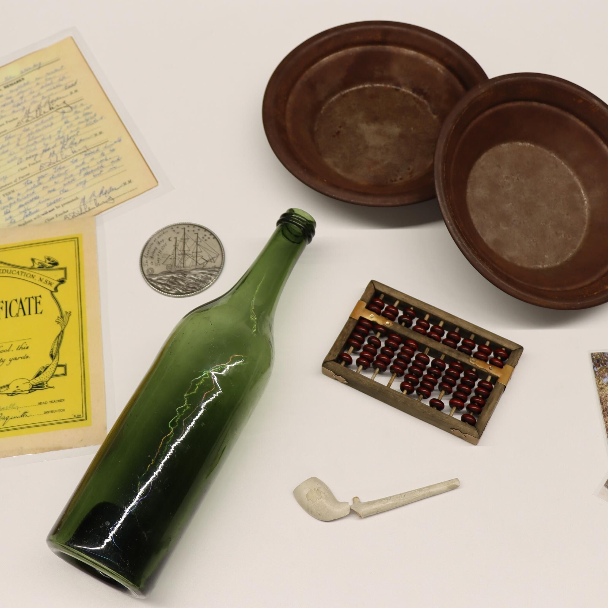 Collection of objects used in education program, including a green glass bottle, a broken clay pipe, metal gold pans, photographs and laminated certificates.  