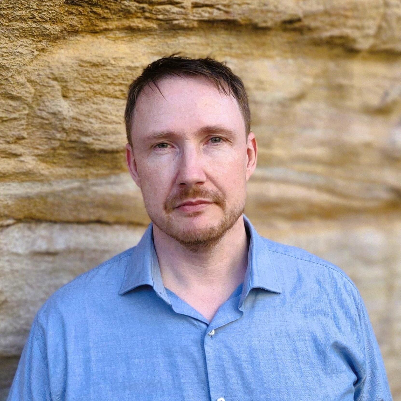 Photograph of a man with light skin, short hair and a blue shirt standing in front of a sandstone wall. 