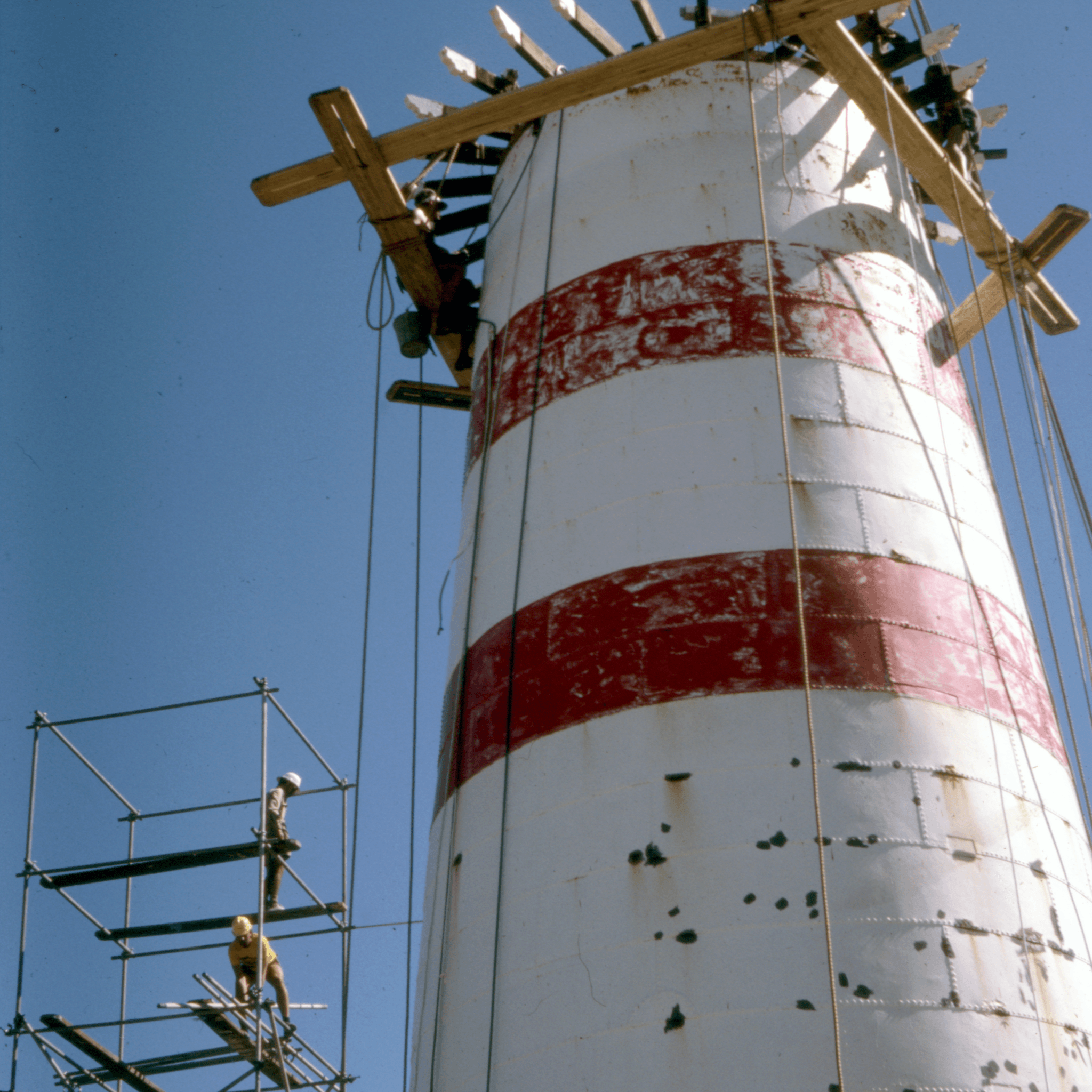 Photograph looking up at a partially dismantled lighthouse which is white, with red stripes, with a blue sky behind.  