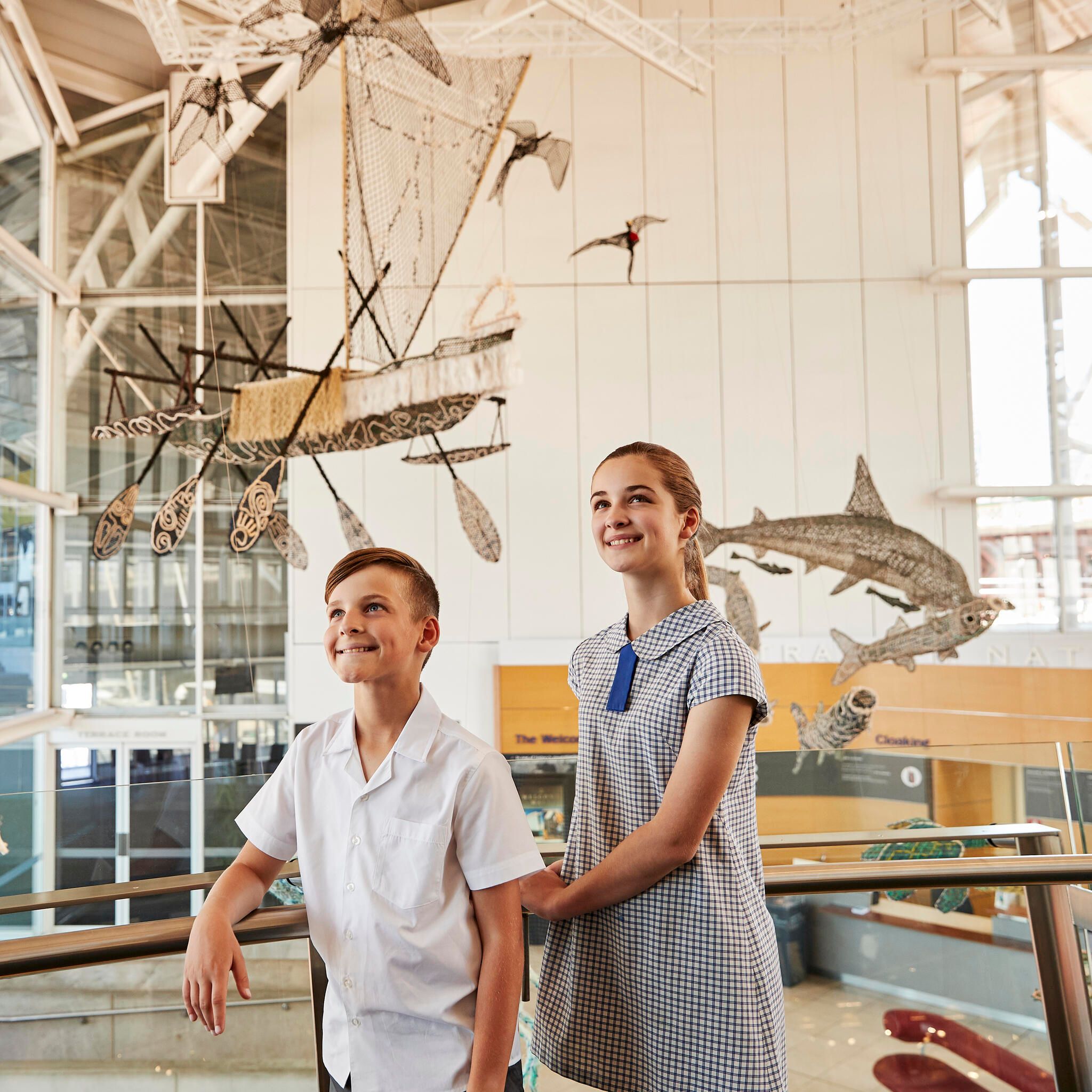 2 School kids exploring the museum, stop to look at the ghost net sculptures suspended in the foyer.