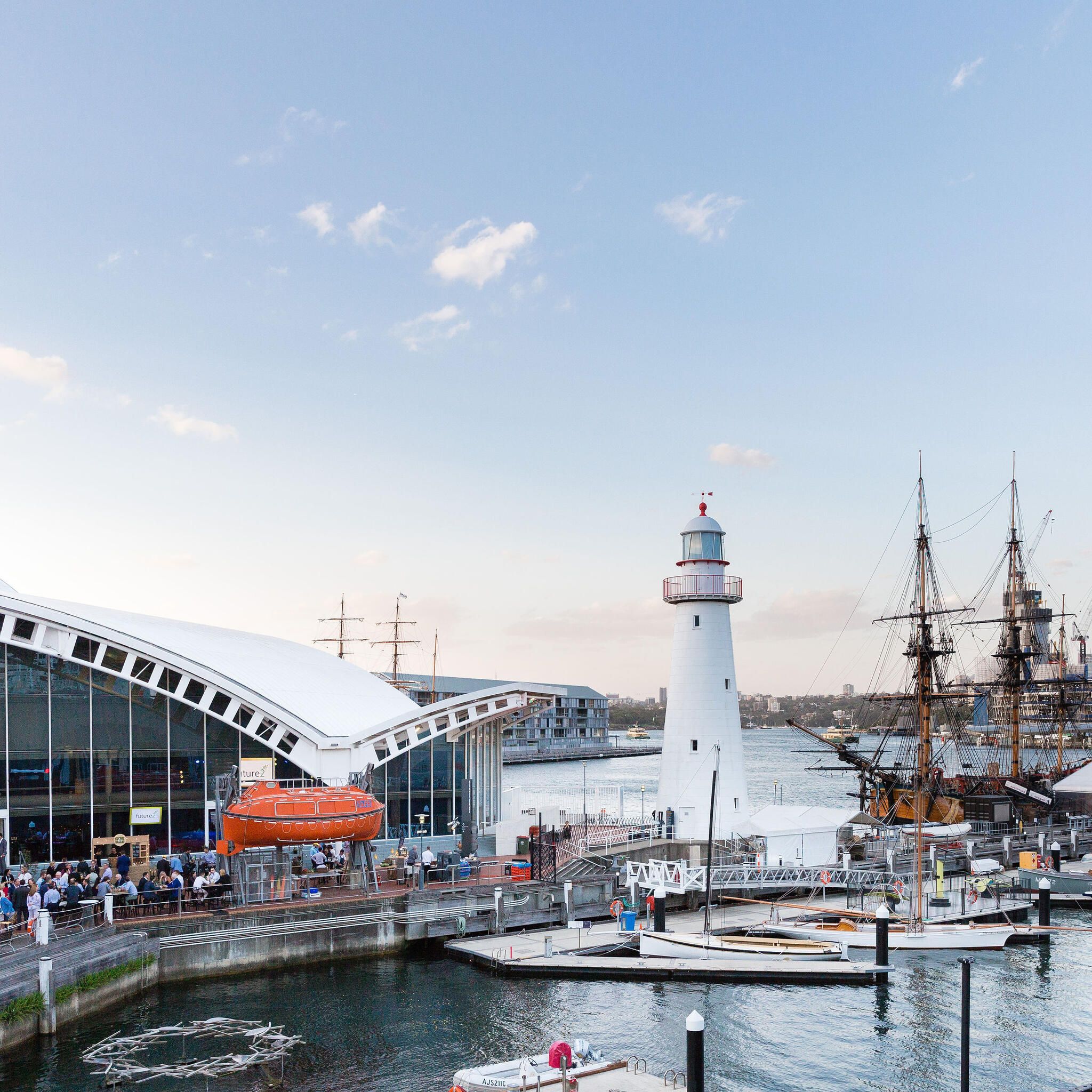 The museum precinct filled with crowds during a corporate event. museum building on the left, with lighthouse and tall ship on the right.