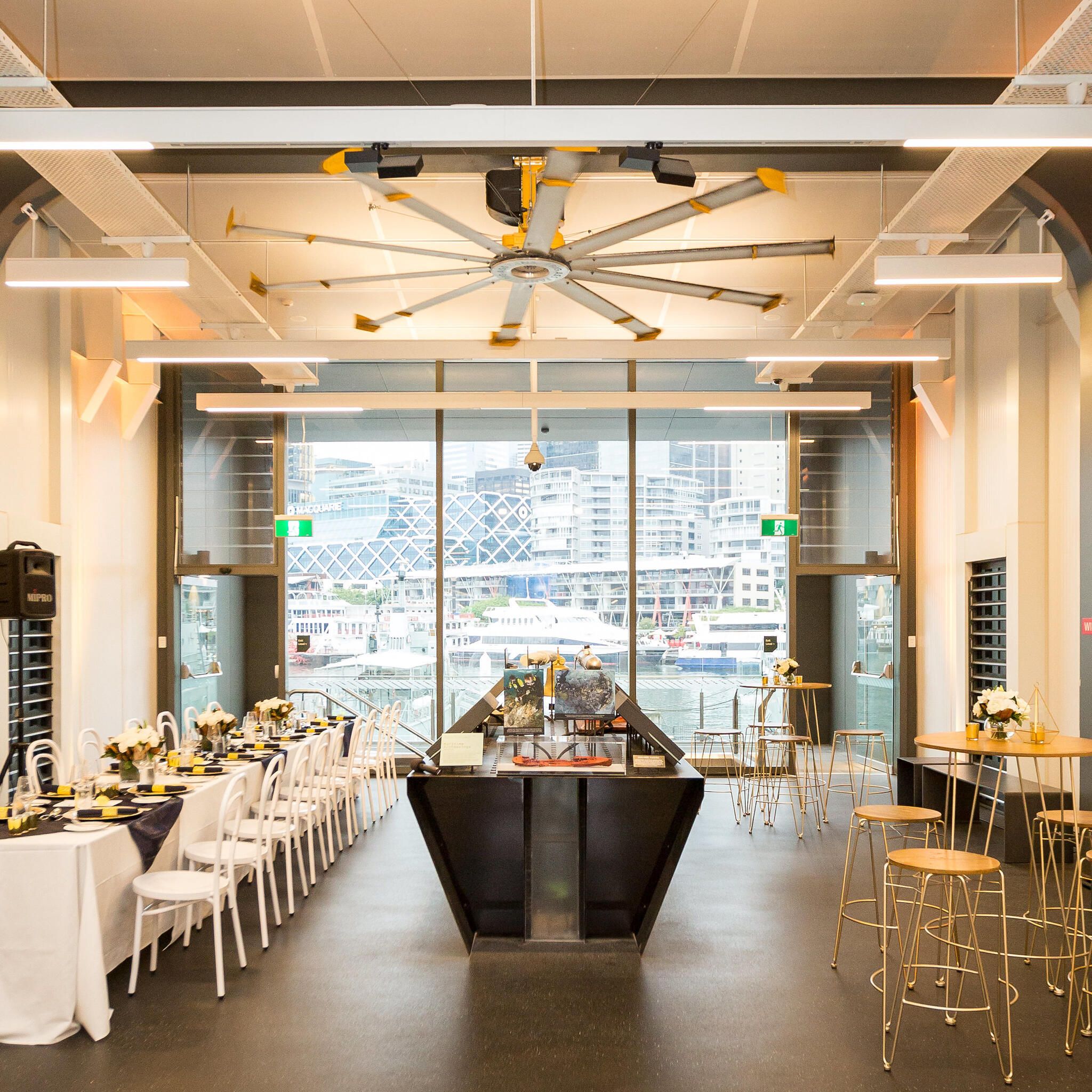 photo of a museum space with a long table set and decorated on the left side, museum interactive displays in the middle and a cocktail table and stools on the right, with a large window to the rear.  