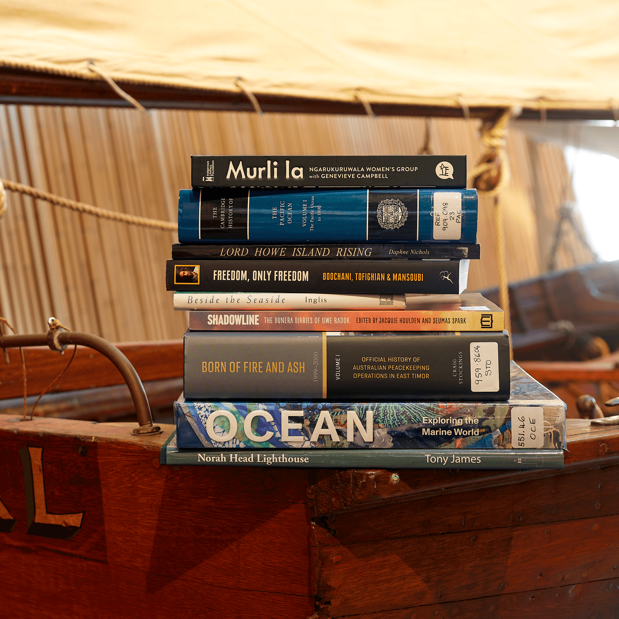 Pile of library books with different coloured covers sitting on te side of a wooden boat. 