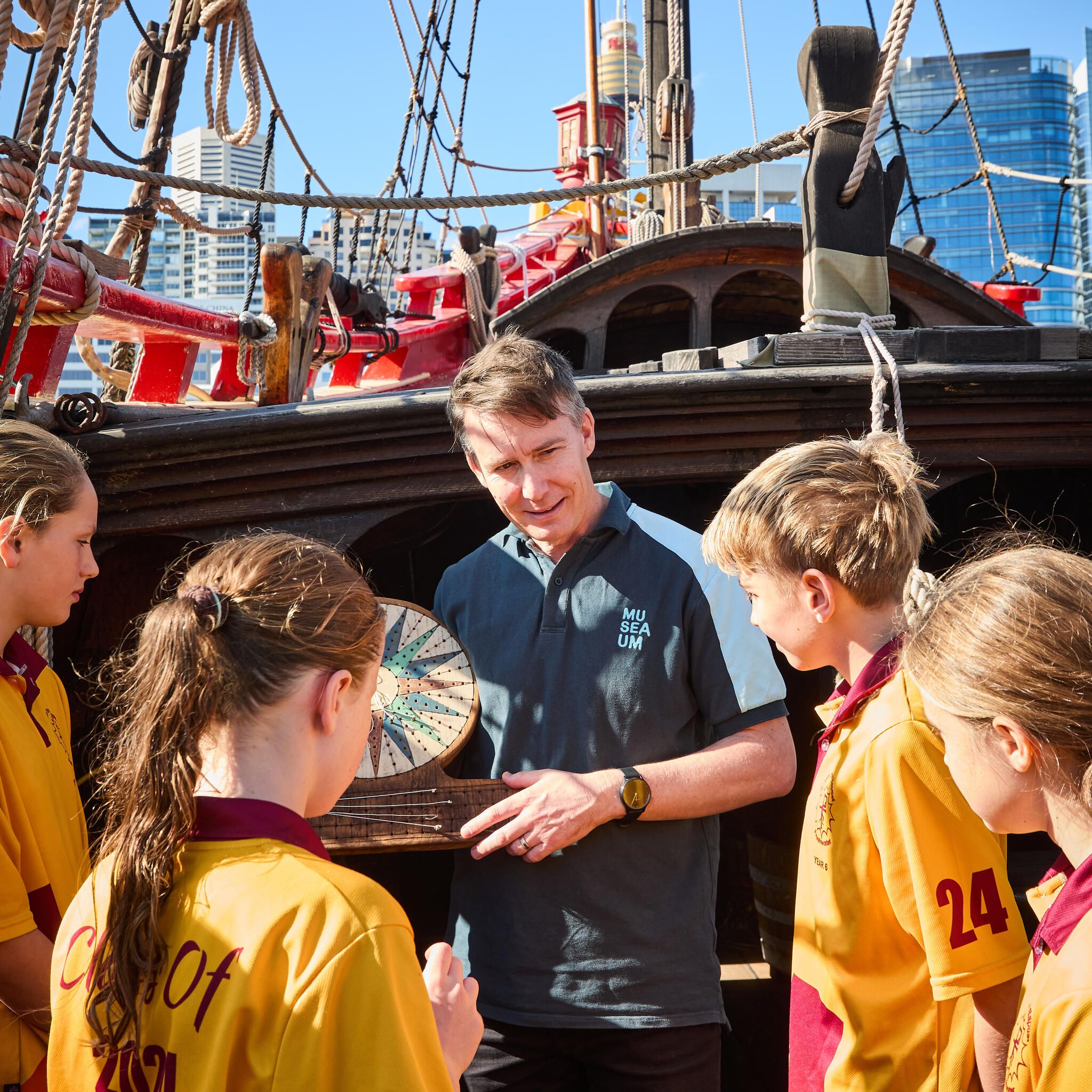 A male museum educator stands with 4 students around him onboard the Duyfken