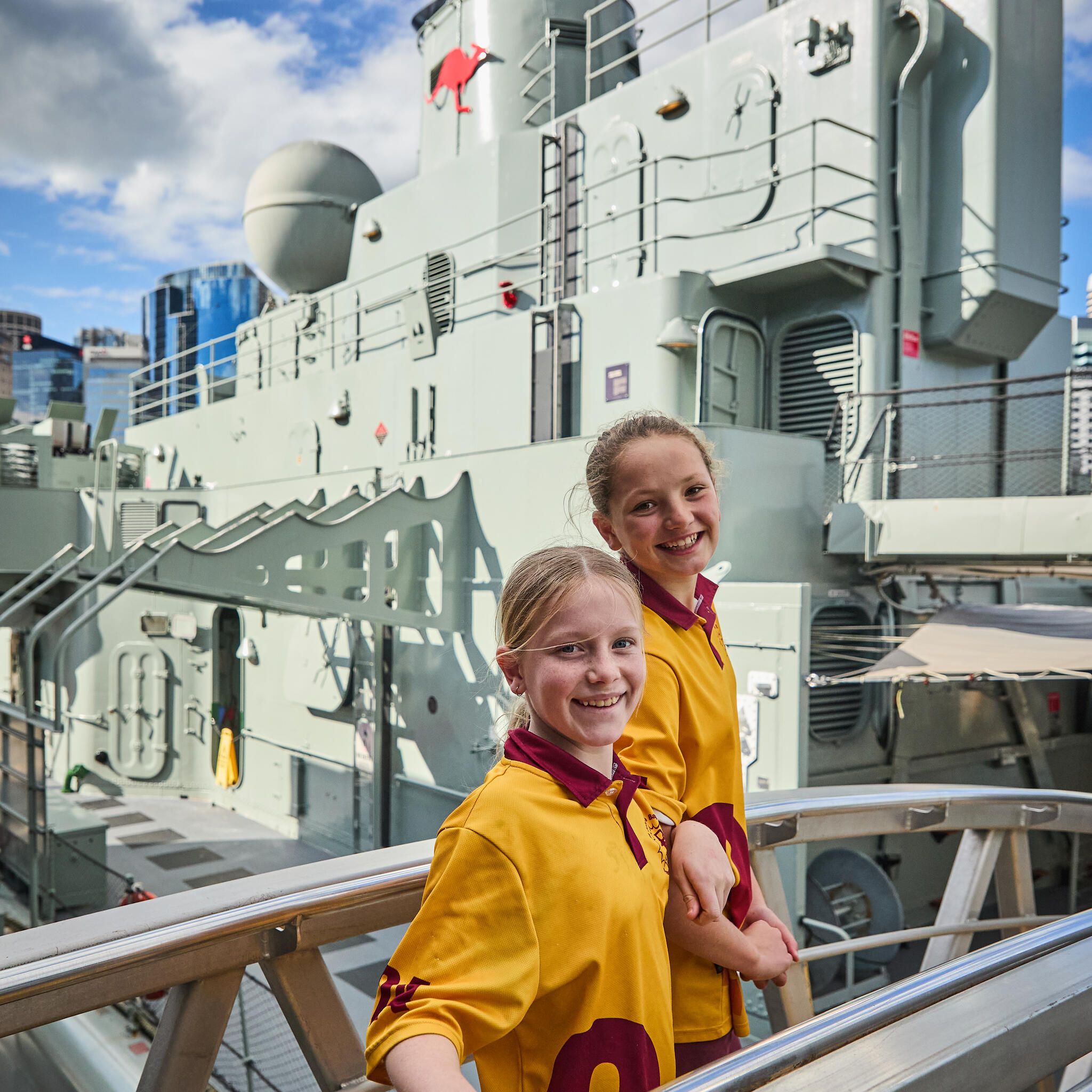 Two firls in a yellow and red school uniform on a gangway with the navy vessel vampire behind them.