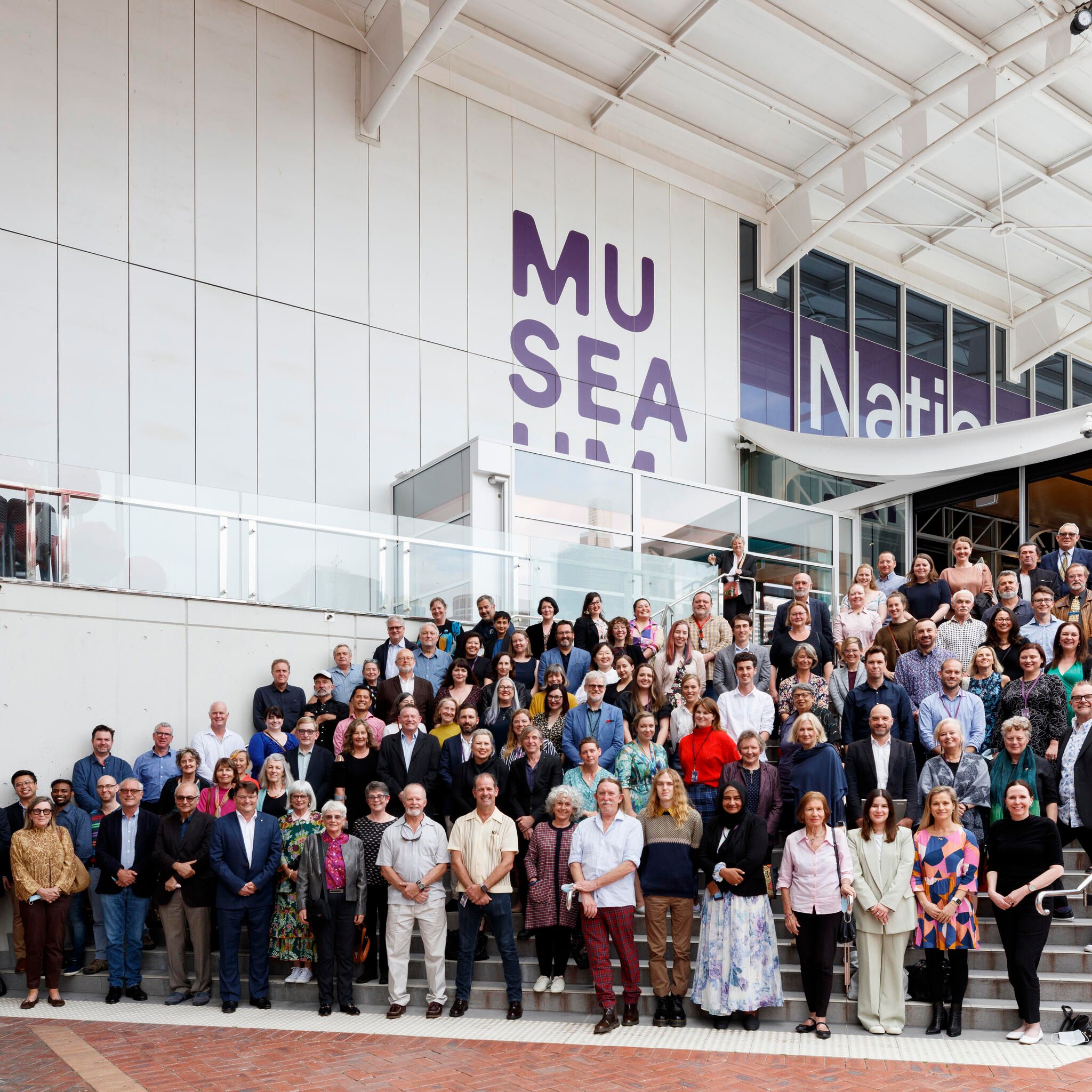 large group of people standing on wide steps in front of a white building with the museum logo on it. 