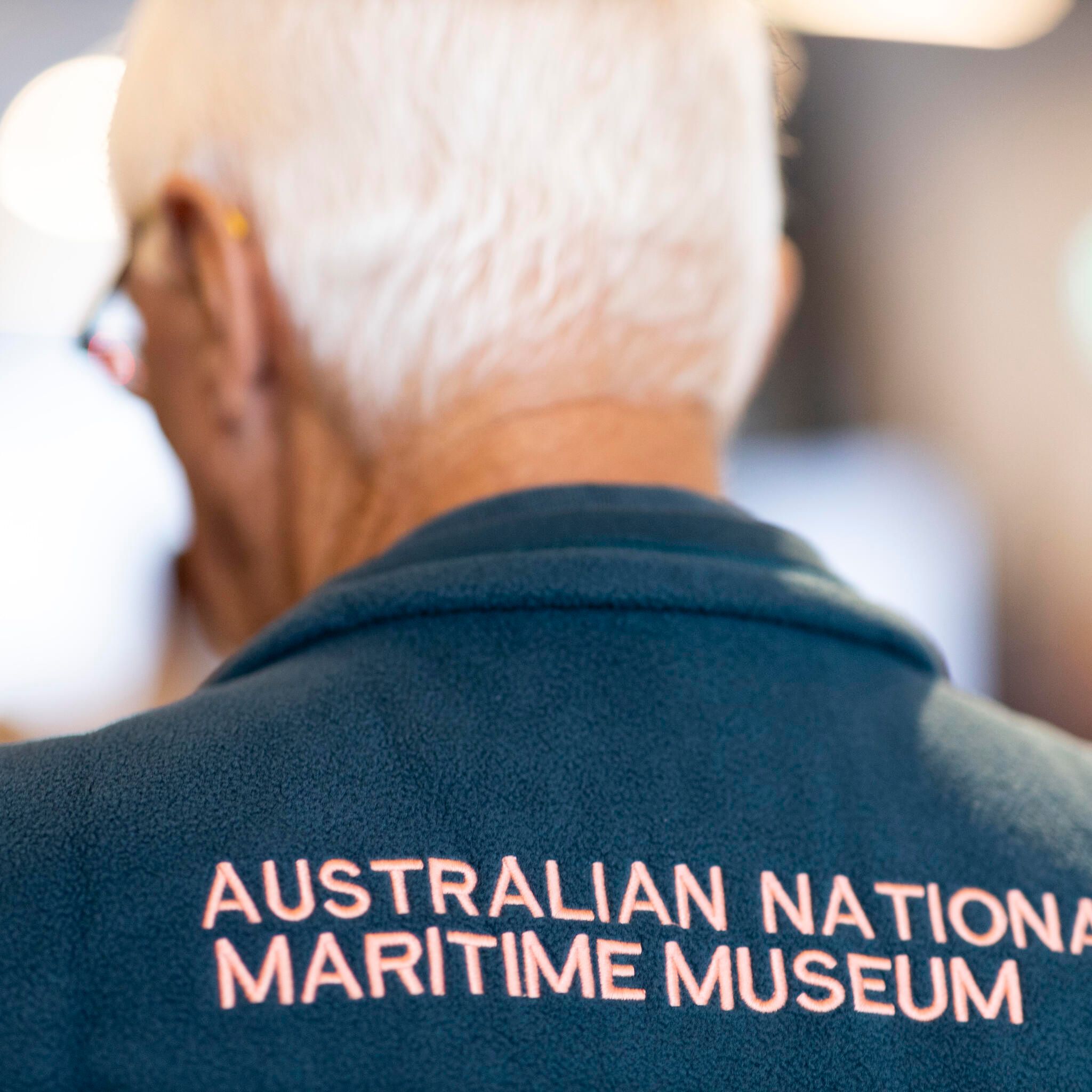 Back of a male museum volunteer with white hair wearing a vest with the text Australian National Maritime Museum. 