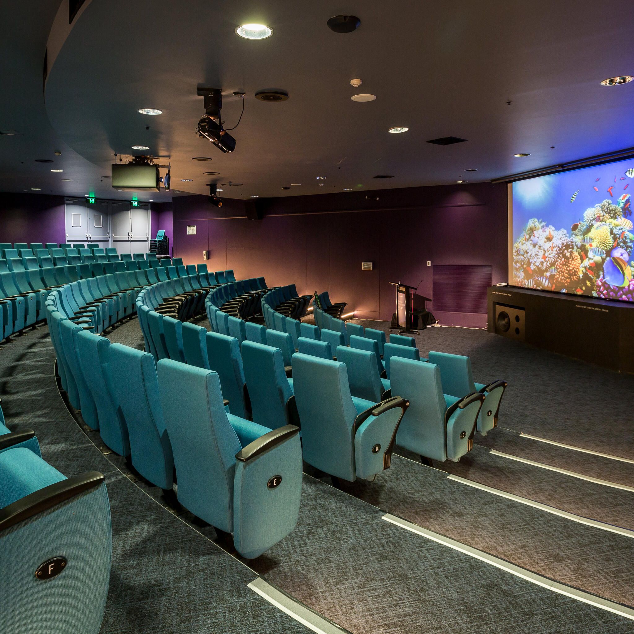 photo of an empty theatre with blue seats and a large screen at the front. 