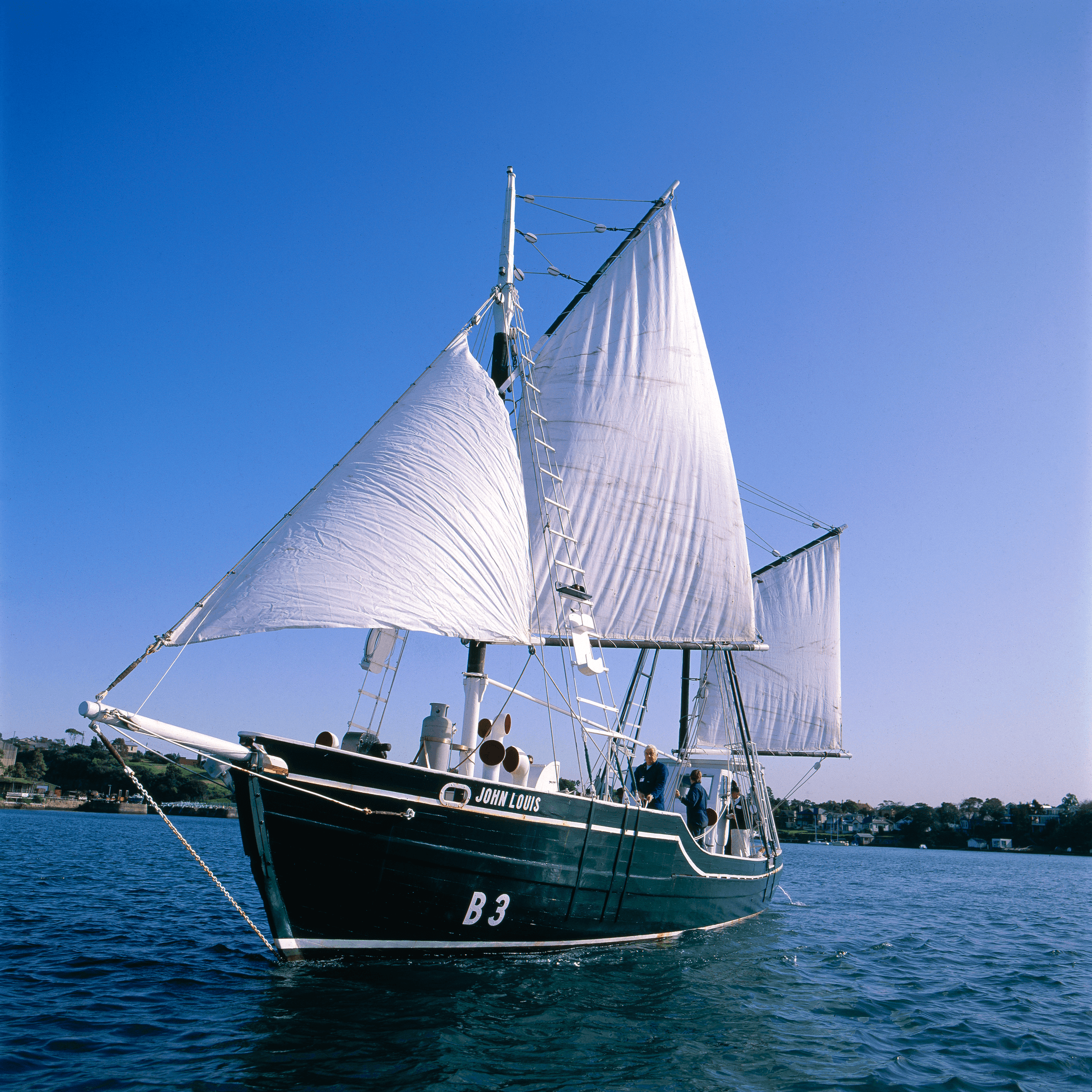 boat with a dark green hull and white sails on blue water with a clear blue sky. 