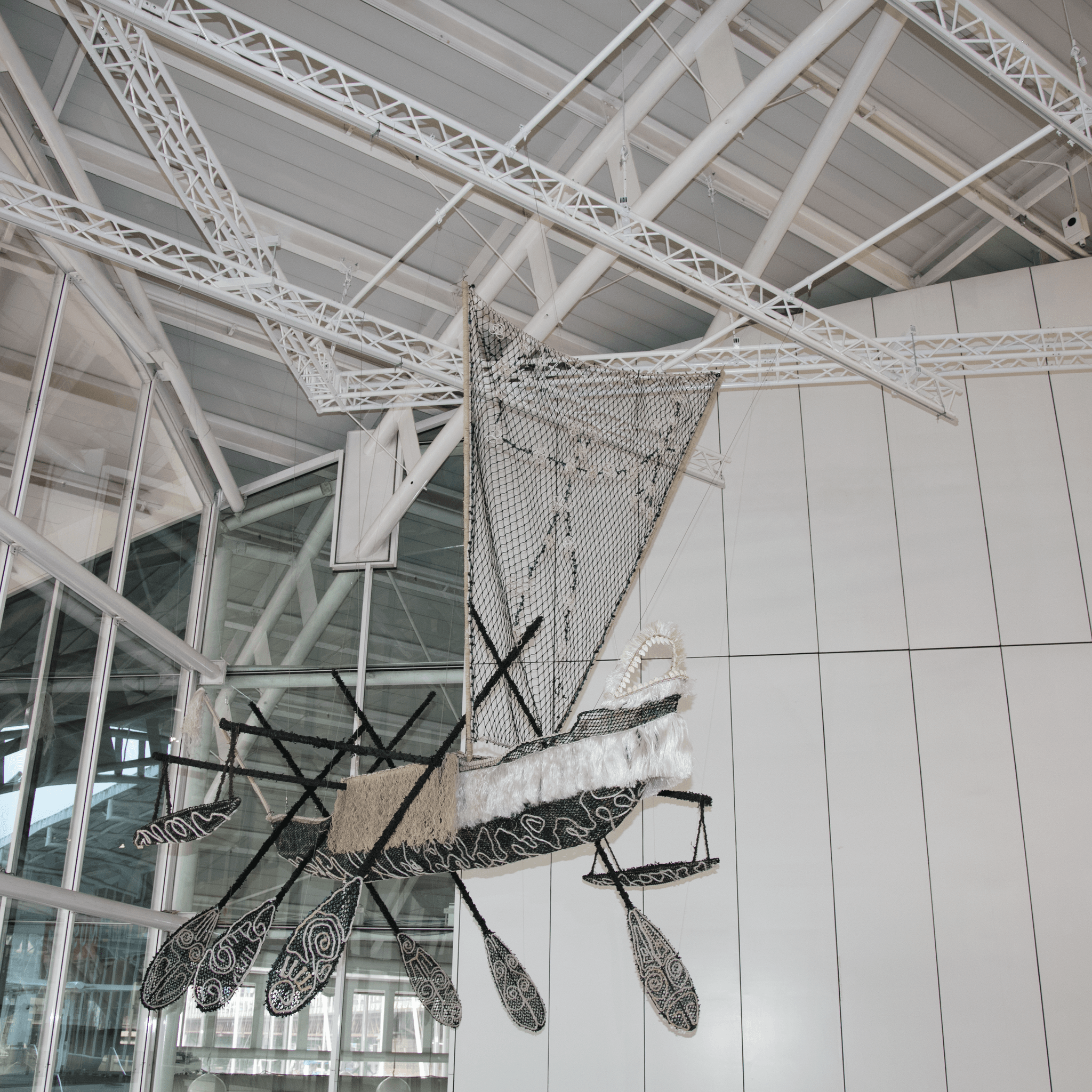 Sculpture in the shape of a boat with a sail and paddles, made of fishing nets and rope hanging from the roof. 