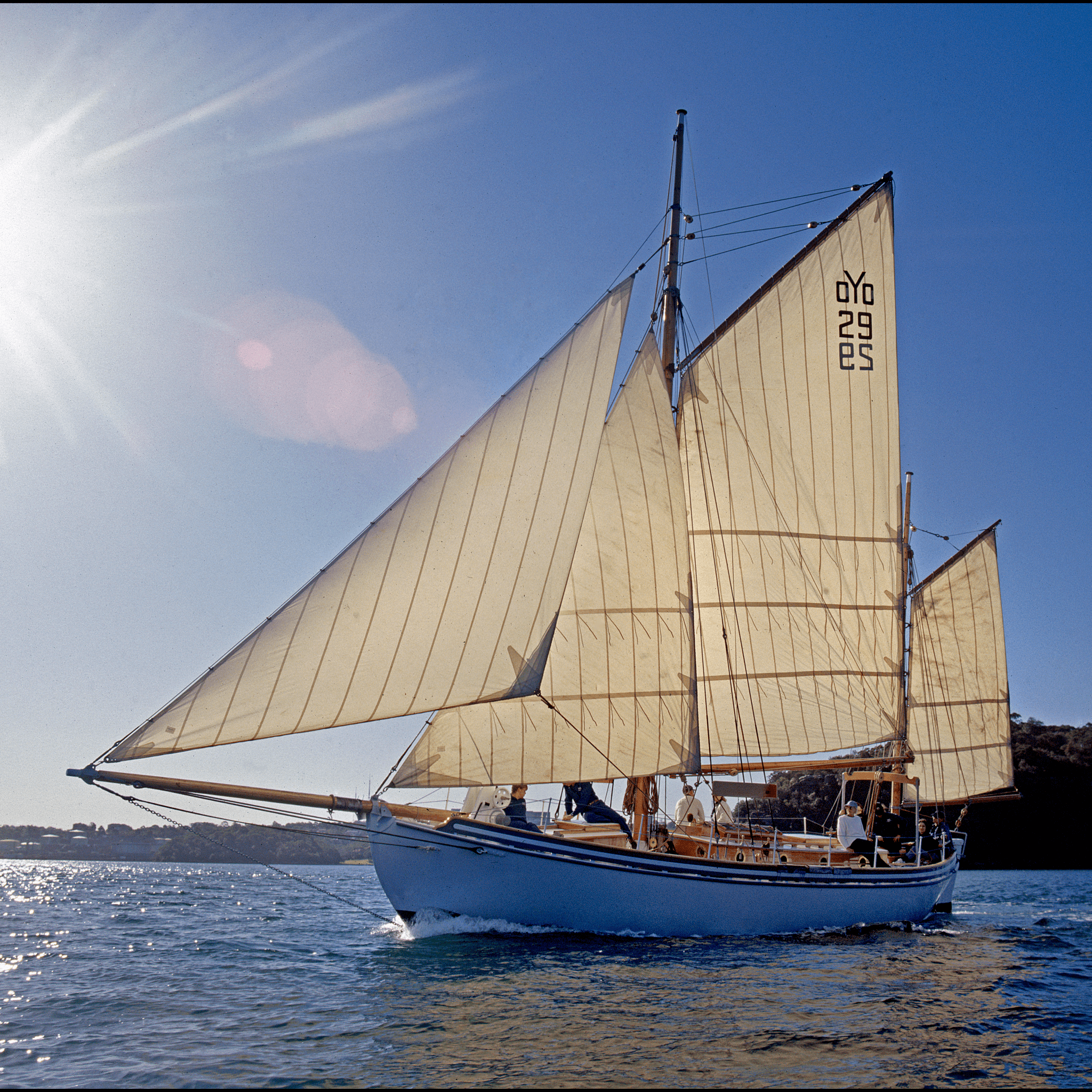 Photo of a small white boat with cream coloured sails, with lens flair from the sun behind it in the clear blue sky. 