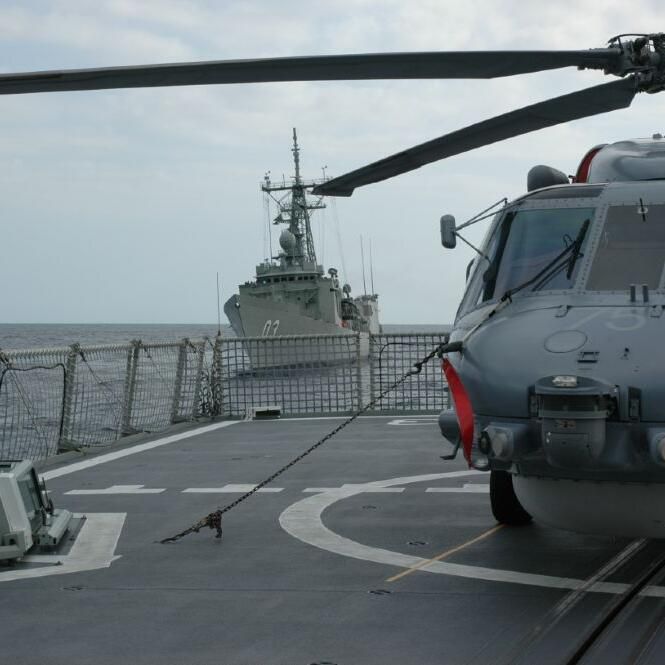 A grey military helicopter on a ship's deck, with another navy ship in the background.